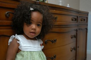 criança negra de cabelos cacheados parada em frente a uma cômoda de madeira