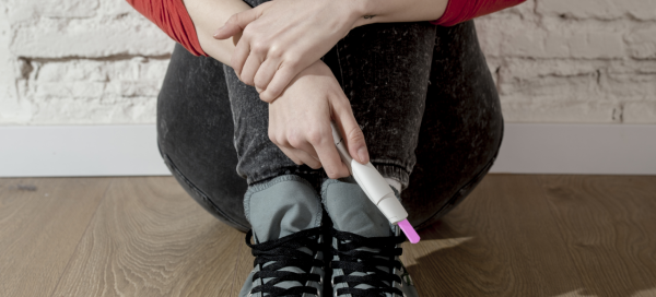 Adolescente de tênis e calça jeans sentada no chão segurando um teste de gravidez