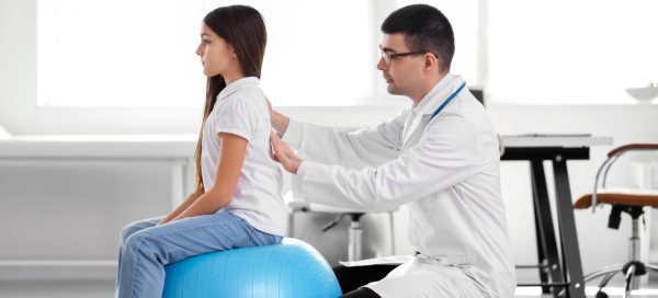 Adolescente de cabelos longos e escuros sentada em uma bola de pilates tem a coluna examinada por um médico