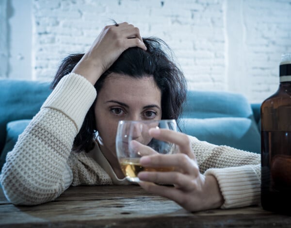 Mulher bêbada sentada no chão enquanto se apoia em uma mesa de madeira e segura um copo de bebida álcoolica