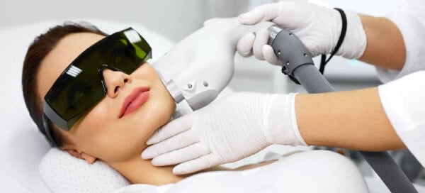 Mulher de óculos escuros de proteção deitada em uma maca em um consultório médico recebendo um tratamento a laser na pele do rosto