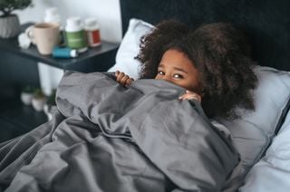Criança negra deitada em uma cama cobrindo o rosto com um edredom cinza