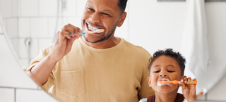 Criança escova os dentes junto com seu pai em frente a um espelho