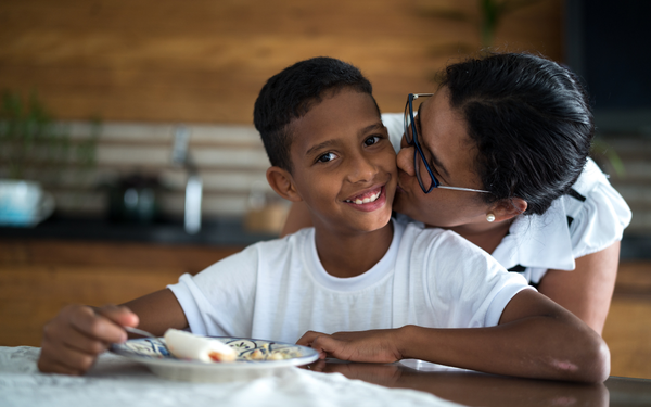 Menino sentado à mesa enquanto a mãe lhe dá um beijo na bochecha