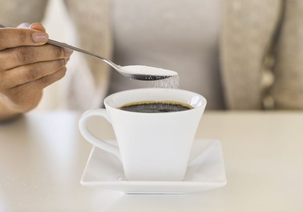 Imagem aproximada de mão feminina colocando uma colher de sopa de açúcar em xícara de café