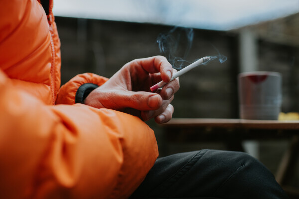 Imagem aproximada de homem segurando um cigarro; ele veste jaqueta laranja e calça preta