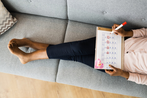Mulher sentada no sofá com as pernas cruzadas, vestindo calça jeans e blusa cor-de-rosa de mangas compridas, e segurando um calendário de ciclo menstrual