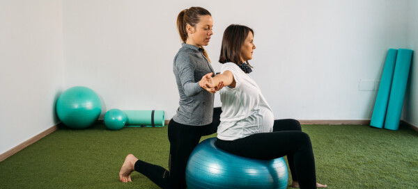 Mulher grávida faz exercício em bola de pilates com auxílio de profissional da saúde