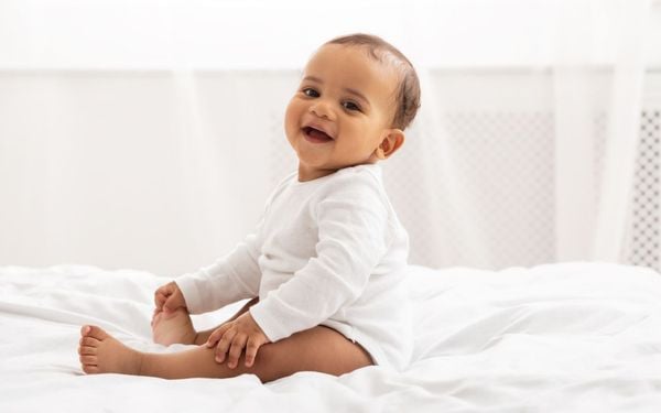 bebê de pele negra sentado na cama olhando para frente e sorrindo