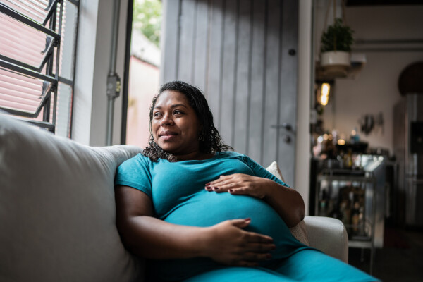 Mulher grávida sentada em sofá enquanto toca a barriga e olha pela janela
