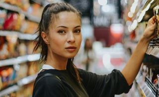 Mulher em um supermercado escolhendo coisas em uma prateleira e olhando para a câmera