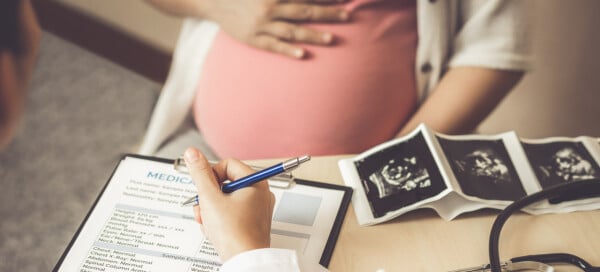 Mulher grávida é atendida por um médico, que anota informações em uma prancheta. Imagens de ultrassom e um estetoscópio estão sobre a mesa