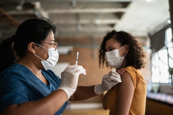 Enfermeira, de pele negra e com vestimenta azul, aplica vacina em adolescente, também de pele negra que veste camiseta amarela