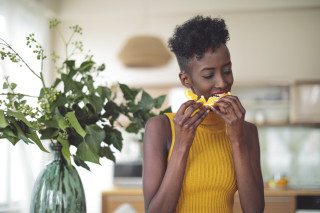 Mulher em pé com uma blusa amarela, comendo laranja com as duas mãos ao lado de um vaso de planta