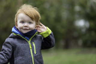 Na imagem, uma criança pequena do sexo masculino posa, com a mão esquerda na cabeça, em um parque
