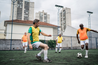 Dois homens maduros, um branco com camiseta verde e colete amarelo, e outro negro, com camiseta bege e colete laranja, jogando futebol