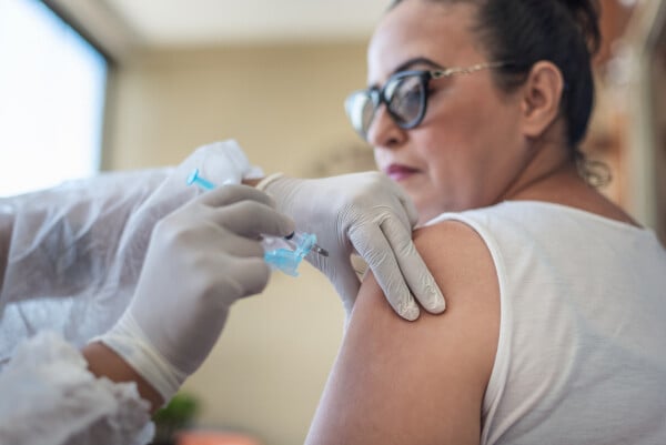 Mulher branca veste regata branca e usa óculos preto enquanto enfermeira aplica vacina da gripe em seu braço