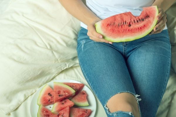 Mulher segurando uma fatia de melancia enquanto está sentada na cama