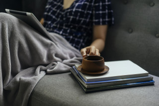 Pessoa sentada em sofá cinza com cobertor nas pernas e tablet nas coxas, com xícara de café em cima de livros ao lado