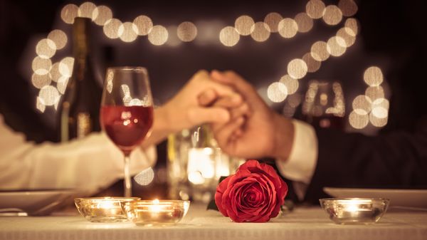 Casal dando as mãos em jantar especial à mesa com taça de vinho e rosa