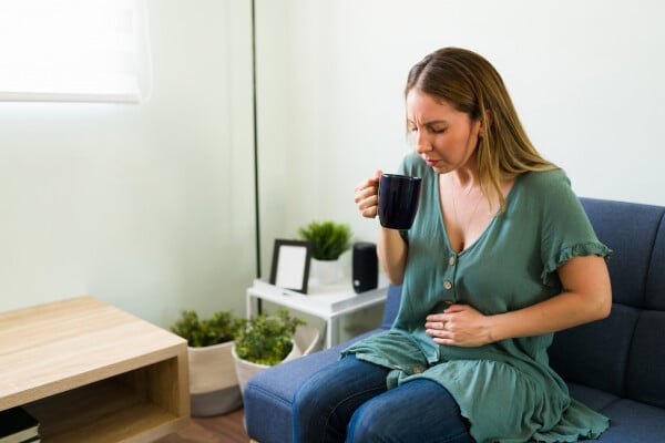 Mulher sentada em um sofá, com uma mão no estômago e outra segurando uma caneca de chá