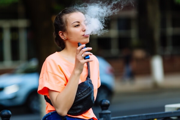Jovem branca, de cabelos pretos presos, camiseta laranja e pochete preta, faz uso de um cigarro eletrônico e solta fumaça pela boca