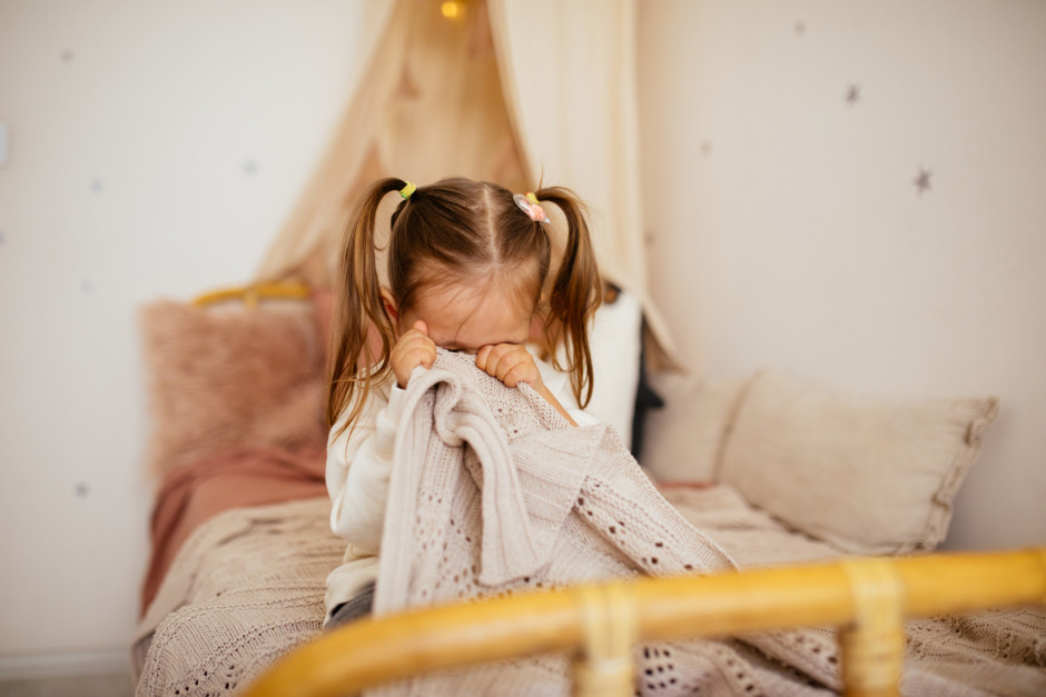 Criança sentada na cama chorando com cobertos no rosto