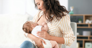 Mulher jovem, branca de cabelos ondulados e escuros, amamentando um bebê branco com cabelos loiros