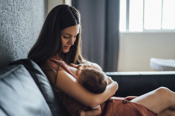 Na imagem, uma mulher sentada em um sofá enquanto amamenta um bebê que está em seu colo