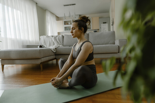 Mulher sentada no chão da sala de casa fazendo uma postura de yoga