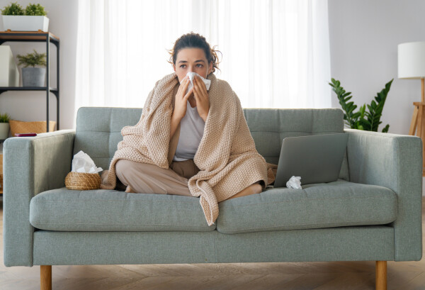 Mulher sentada no sofá, coberta por uma manta, assoando o nariz