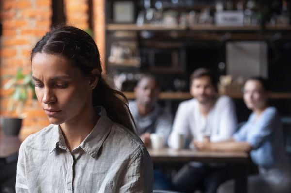 Mulher com feição triste; atrás dela, três pessoas desfocadas, interagindo