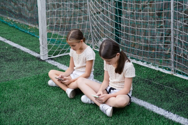 Duas meninas de aproximadamente 10 anos sentadas em um campo de futebol mexendo no celular