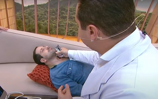 apresentador Paulo Mathias durante o programa Chega Mais, sentado em um sofá enquanto um médico realiza um ultrassom em sua tireoide