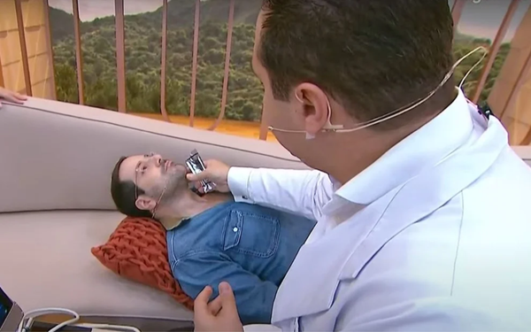 apresentador Paulo Mathias durante o programa Chega Mais, sentado em um sofá enquanto um médico realiza um ultrassom em sua tireoide
