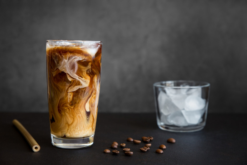 Café gelado em vidro alto com creme, recipiente com gelo e grãos de café em fundo escuro.