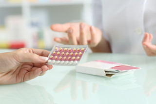 Imagem aproximada de mão feminina, pele branca, segurando uma cartela de pílula anticoncepcional; ao fundo, par de mãos femininas, de pele branca também.