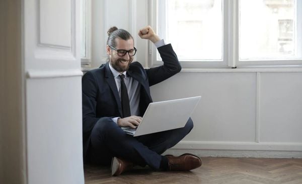 Homem sentado no chão com um notebook no colo comemorando