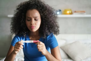 Mulher negra, com cabelos crespos, vestindo uma camiseta de manga curta azul e segurando um teste de gravidez. Sua expressão facial é de preocupação.