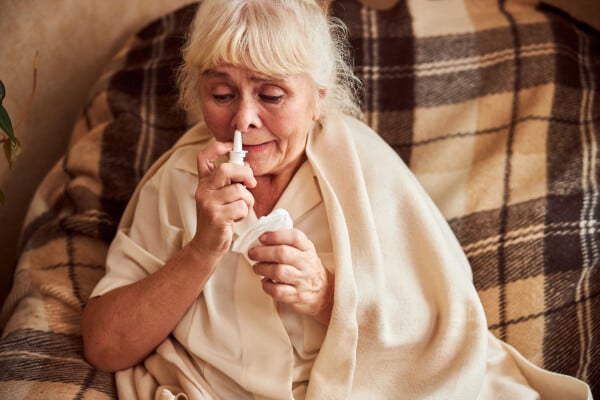 mulher idosa sentada em sofá com um pano ao redor dos ombros usando um spray nasal