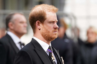 Príncipe Harry veste terno preto, camisa branca e gravata preta; aparece de perfil na foto