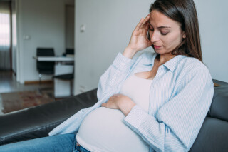 Mulher grávida sentada em sofá com uma mão na barriga e outra na testa