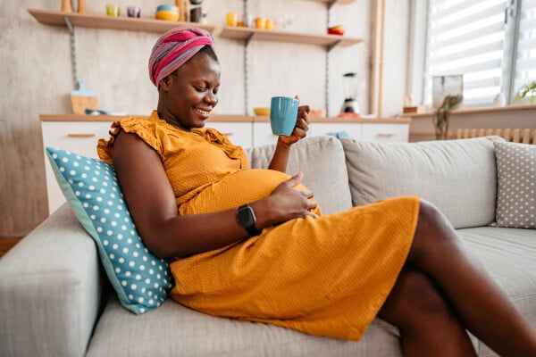 Mulher negra grávida sentada em um sofá tomando chá enquanto passa a mão pela barriga