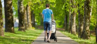 Homem veste bermuda jeans e camiseta azul enquanto empurra um carrinho de bebê em meio a um parque cheio de árvores