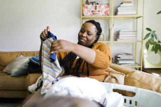 Mulher sentada no sofá pegando roupa de bebê de uma cesta de roupa