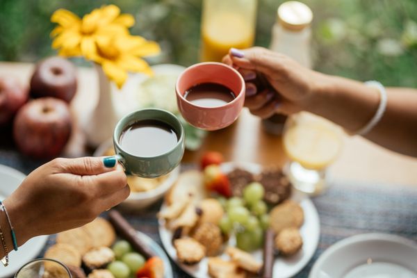 Imagem aproximada de duas mãos brindando com xícaras de café. Ao fundo, em desfoque, uma mesa com um café da manhã variado