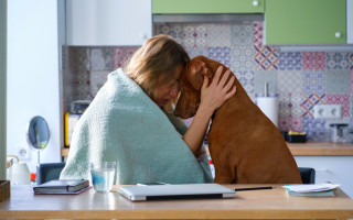 Mulher sentada em mesa enrolada em uma manta azul enquanto abraça um cachorro