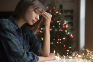 Mulher de blusa azul sentada apoiando o cotovelo na mesa com uma árvore de natal no fundo