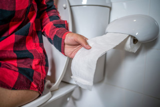 Mulher sentada em vaso sanitário enquanto puxa um pedaço de papel higiênico para se limpar