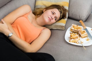 Na imagem, uma mulher está deitada em um sofá, com as mãos sob o estomago, enquanto olha um prato com pedaços de torta